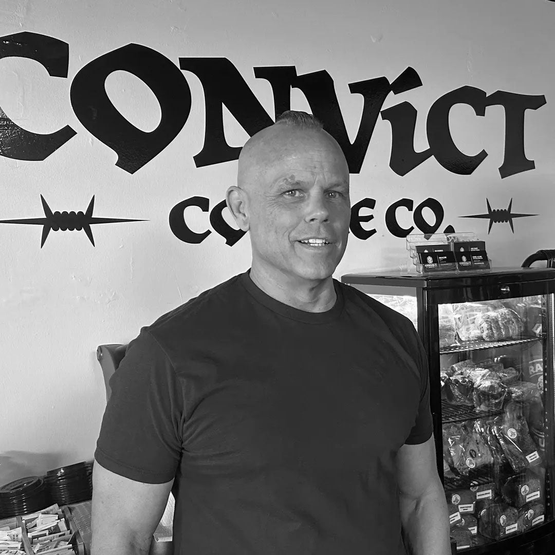 A black and white photo of Dan Klehm standing in front of a wall with a large 'CONVICT COFFEE CO.' sign and a glass display case of packaged goods.
