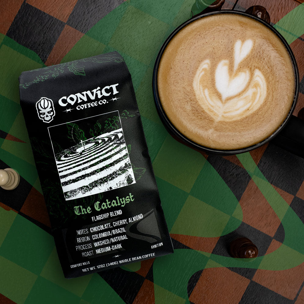 A bag of Convict Coffee Co. 'The Catalyst' espresso roast coffee is shown next to a black mug filled with latte art coffee, set on a chessboard.