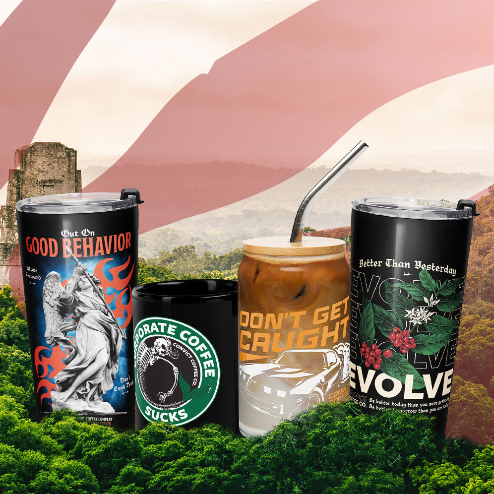 Four novelty tumbler cups arranged together in a stylized outdoor scene with a mountain landscape and red-and-white flag background. The cups include graphic designs: one with a statue and flames reading 'OUT ON GOOD BEHAVIOR,' one black with a skull logo and the words 'CONVICT COFFEE SUCKS,' one amber drink with orange text reading 'DON'T GET CAUGHT,' and one black cup with white text and botanical artwork reading 'Better Than Yesterday.