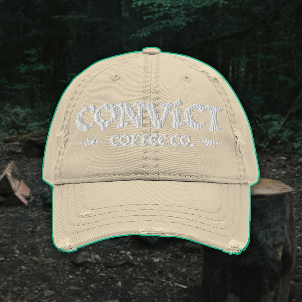 Khaki Distressed Dad Hat with 'Convict Coffee Co.' logo white embroidery on a Forest Campsite background with cut wood