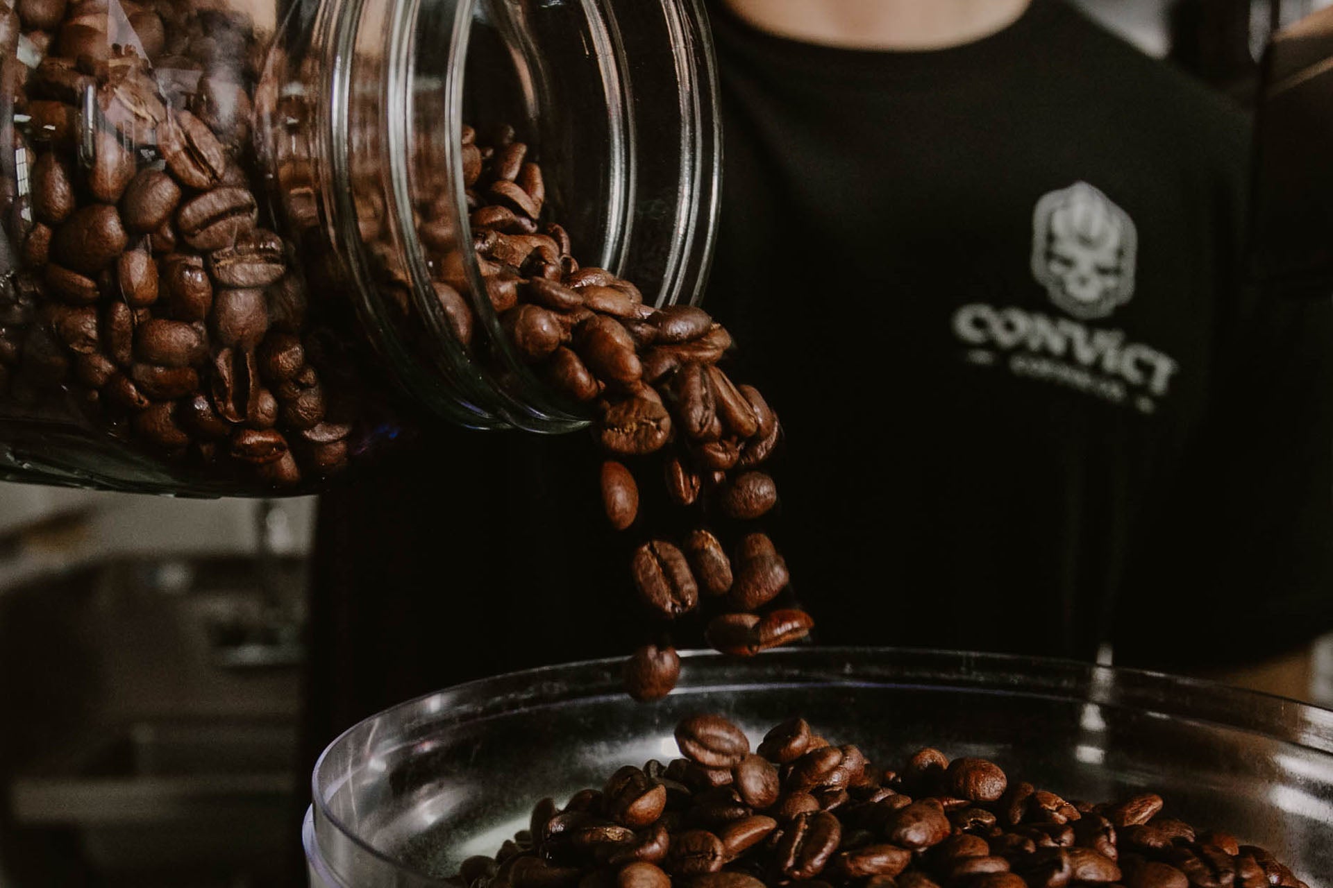 Coffee beans being poured from a jar into a grinder hopper with 'Convict Coffee Co.' shirt in background
