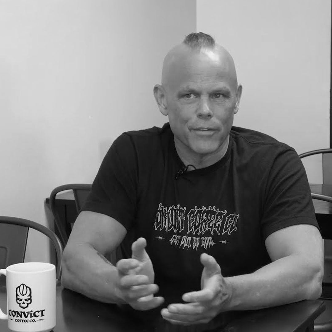 A black and white photo of Dan Klehm sitting at a table, wearing a black graphic t-shirt. A white coffee mug with the 'Convict Coffee Co.