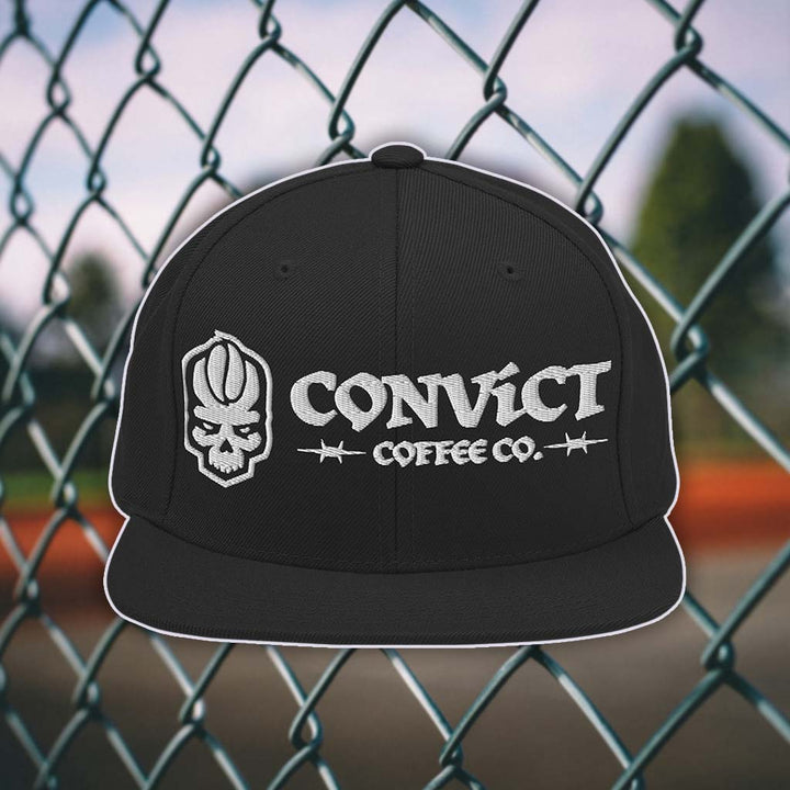 Black snapback flat bill with 'Convict Coffee Co.' logo white embroidery on a chain-link fence background